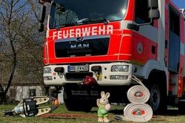 Feuerwehrauto, davor ein Deko Osterhase, die Rettungsschere mit einem Ei, grünes Gras und blauer Himmel. Darauf der Schriftzug.