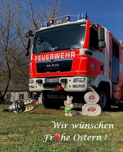 Feuerwehrauto, davor ein Deko Osterhase, die Rettungsschere mit einem Ei, grünes Gras und blauer Himmel. Darauf der Schriftzug.