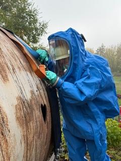 Ein Mann in blauem Schutzanzügen am Chemiefass von der Seite