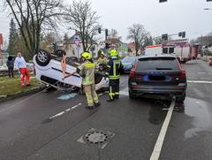 Weißer PKW liegt auf dem Dach. Feuerwehrmänner stehen davor.