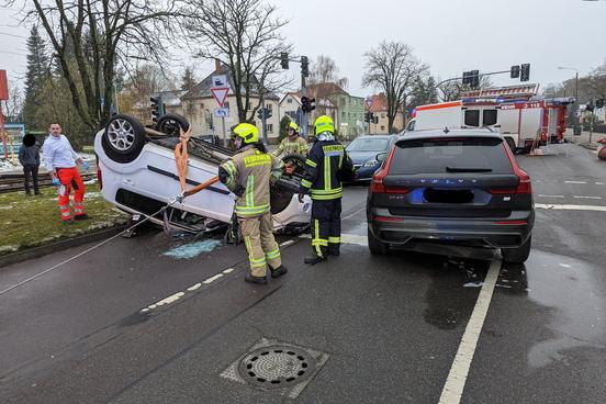 Weißer PKW liegt auf dem Dach. Feuerwehrmänner stehen davor.