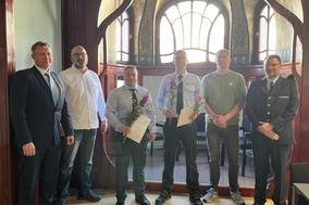 Sechs Männer in einem Büro, Zwei haben eine Urkunde und Blumen in der Hand.