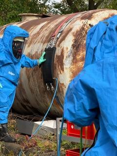 zwei Männer in blauen Schutzanzügen am Chemiefass