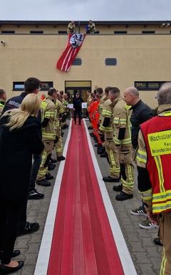 Zwei Feuerwehrmänner stehen auf Terrasse mit Wassereimern in der Hand. Darunter stehen Feuerwehrmänner Spalier.