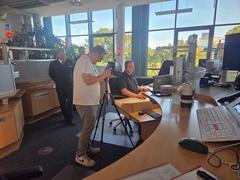 Mitarbeiter in der Leitstelle am Tisch, davor ein Mann, der Aufnahmen mach und im Hintergrund weitere Mitarbeiter.