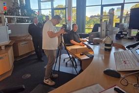 Mitarbeiter in der Leitstelle am Tisch, davor ein Mann, der Aufnahmen mach und im Hintergrund weitere Mitarbeiter.
