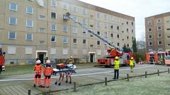 Wohnblock mit mehreren Fahrzeugen der Feuerwehr davor. Rauch tritt aus dem Fenster.