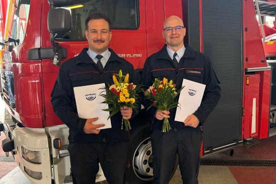 Zwei Kollegen mit Blumenstrauß und Zeugnissen vor einem Feuerwehrauto
