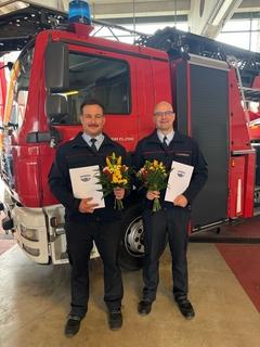 Zwei Kollegen mit Blumenstrauß und Zeugnissen vor einem Feuerwehrauto