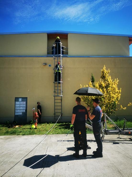 2 Feuerwehrmänner mit einem sonnen schirm im Vordergrund. Im Hintergrund steigt einer eine Notleiter hinauf