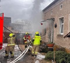 Drei Feuerwehrmänner am Hauseingang. Aus dem Haus tritt Rauch aus.