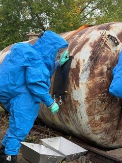 Ein Mann in blauem Schutzanzügen am Chemiefass von der anderen Seite