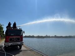 2 Männer stehen auf einem Feuerwehrauto am Silokanal. Sie geben Wasser aus dem Schlauch.
