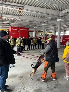 Viele Feuerwehrmänner stehen in der Fahrzeughalle. Die Hundetrainerin erklärt am Hund.