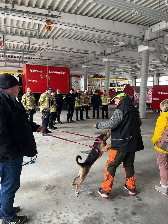 Viele Feuerwehrmänner stehen in der Fahrzeughalle. Die Hundetrainerin erklärt am Hund.