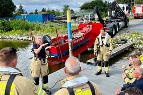 Feuerwehrfrau erklärt Feuerwehrmännern am Steg etwas. Im Hintergrund ist das Rettungsboot