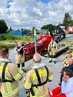 Feuerwehrfrau erklärt Feuerwehrmännern am Steg etwas. Im Hintergrund ist das Rettungsboot