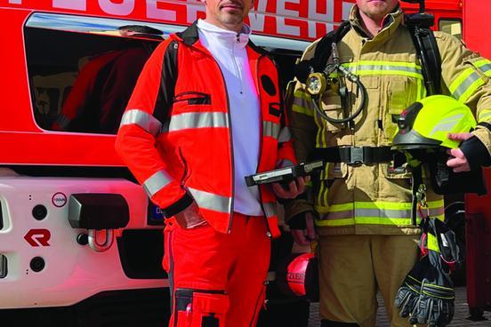 Vor einem Feuerwehrfahrzeug stehen zwei Männer. Einer trägt Rettungsdienstbekleidung und der andere ist Feuerwehrmann