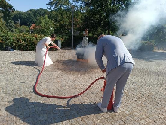 Löschversuch der frisch vermählten. Er an der Kübelspritze, Sie muss löschen