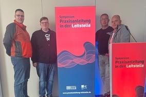 Vier Kollegen am Werbebanner zum Symposium