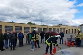 Mehrere Menschen stehen und schauen, wie eine Person mit deinem Feuerlöscher einen Brand in einer Feuerschale löscht.
