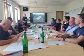 Ein großer Tisch mit Mitarbeitern daran. Im Hintergrund ein Smartboard.