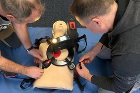 Zwei Männer üben an einer Übungspuppe. Sie haben dafür ein spezielles Gerät.