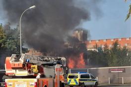 Drehleiter vor Mauer, dahinter Feuer und schwarzer Rauch.