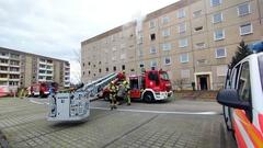 Wohnblock mit 4 Aufgängen. Davor steht die Drehleiter. Aus dem Fenster tritt Rauch