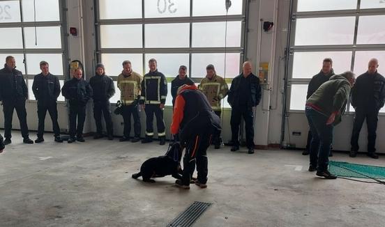 Feuerwehrmänner in der Fahrzeughalle. Zwei Trainer halten ihre Hunde an der Leine