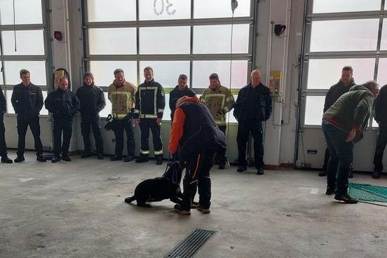 Feuerwehrmänner in der Fahrzeughalle. Zwei Trainer halten ihre Hunde an der Leine