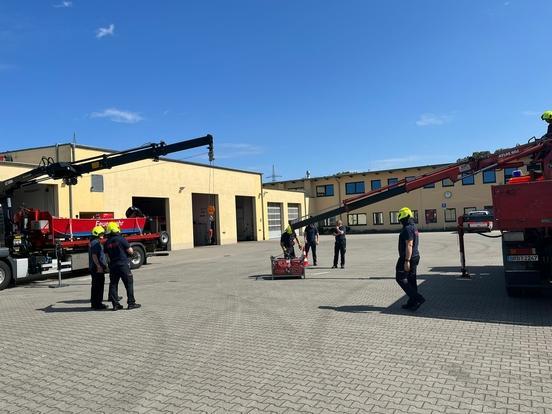 Kran der Feuerwehr hebt Korb mit Schläuche. Mehrere Feuerwehrmänner stehen mit Helmen auf dem Kopf und schauen zu.