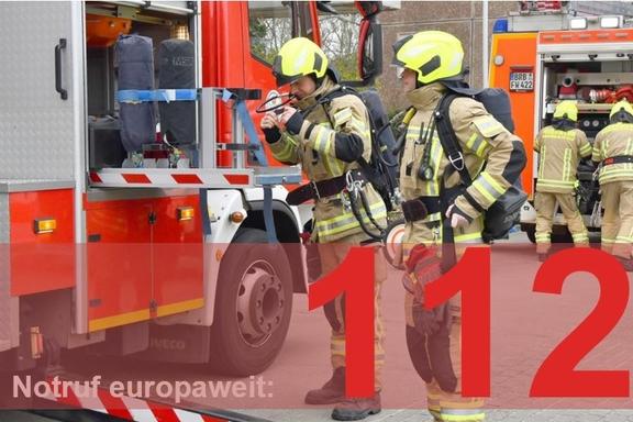 Vier Feuerwehrmänner am Fahrzeug. Im Vordergrund steht Notruf europaweit 112