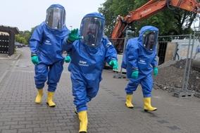 3 Männer in blauen Schutzanzügen und gelben Stiefeln laufen auf dem Hof