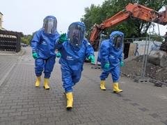 3 Männer in blauen Schutzanzügen und gelben Stiefeln laufen auf dem Hof