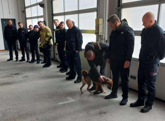 Feuerwehrmänner in der Fahrzeughalle. Sie stehen in einer Reihe. Einer streichelt einen kleinen Hund.