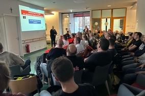 Sitzende Menschen in einem großen Raum mit Leinwand